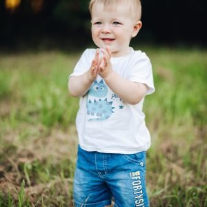 Stock photo portrait in t-shirt and denim shorts clapping hands and smiling standing on green lawn in park. Bokeh background. Cheerful baby boy standing on grass with hands clapping.